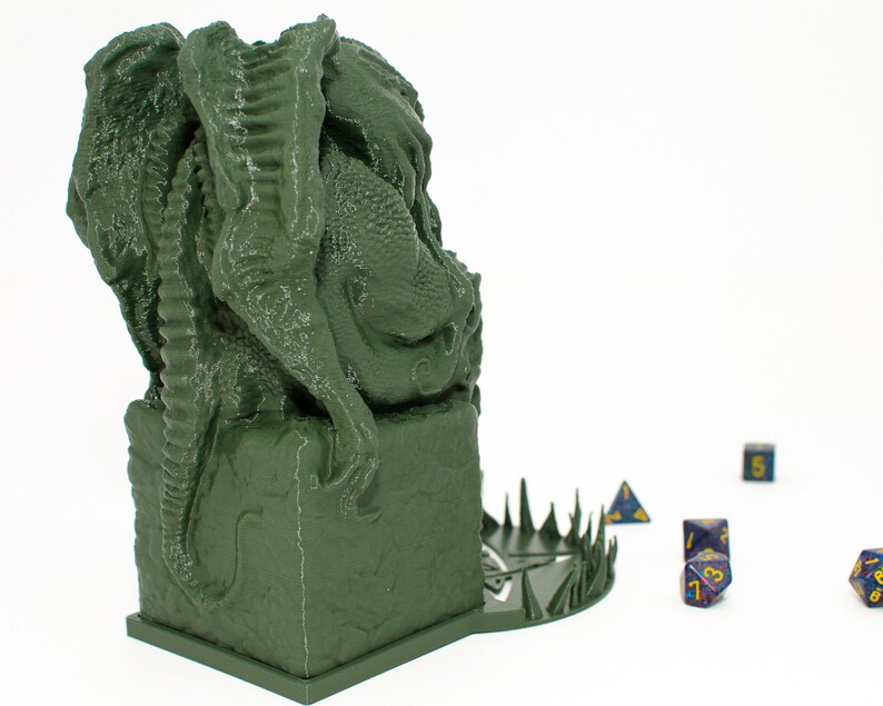 May include: Green 3D printed dragon statue with a base that has a circular design with spikes around the edge. The dragon is sitting on a square base. There are several blue dice on the table in front of the dragon.