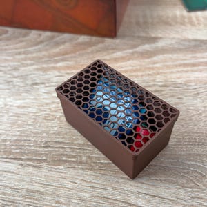 K&ouml;nnte beinhalten: Eine braune rechteckige Box mit einem Wabenmuster auf dem Deckel. Die Box enth&auml;lt farbenfrohe Gegenst&auml;nde, m&ouml;glicherweise W&uuml;rfel oder kleine Spielsteine. Die Box besteht aus einem festen Material und steht auf einer Holzoberfl&auml;che.
