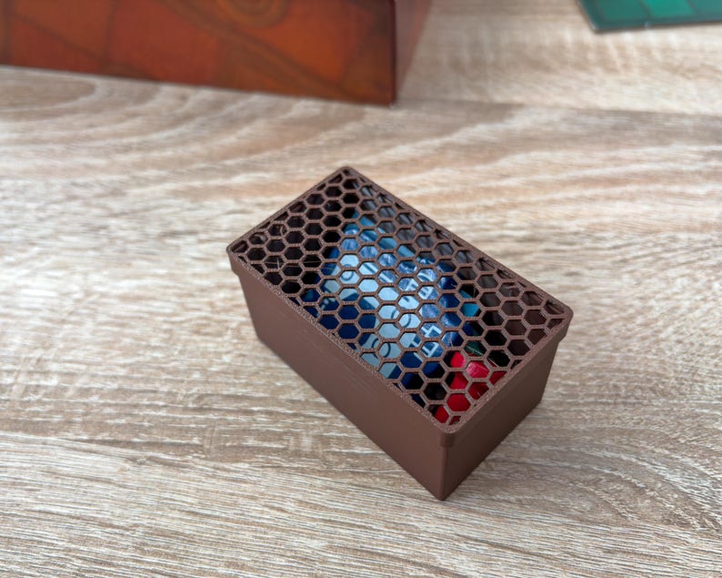 K&ouml;nnte beinhalten: Eine braune rechteckige Box mit einem Wabenmuster auf dem Deckel. Die Box enth&auml;lt farbenfrohe Gegenst&auml;nde, m&ouml;glicherweise W&uuml;rfel oder kleine Spielsteine. Die Box besteht aus einem festen Material und steht auf einer Holzoberfl&auml;che.
