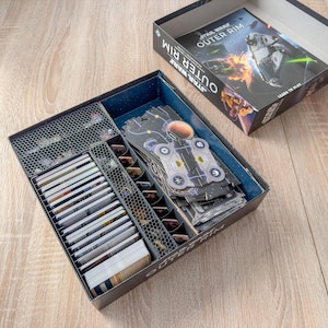 May include: A black board game box labeled "Star Wars Outer Rim" is open, revealing organized game components. Inside are cards, tokens, and game pieces, neatly arranged in compartments. The box is on a wooden surface.