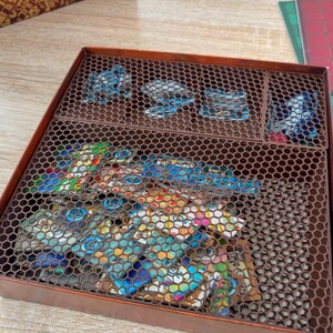 K&ouml;nnte beinhalten: Eine braune Spielbox mit sechseckigem Gitterdeckel, die Spielsteine enth&auml;lt. Die Box ist in F&auml;cher unterteilt, mit bunten Spielkarten und blauen Spielmarken. Die Box steht auf einer Holzoberfl&auml;che.