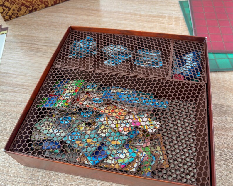 K&ouml;nnte beinhalten: Eine braune Spielbox mit sechseckigem Gitterdeckel, die Spielsteine enth&auml;lt. Die Box ist in F&auml;cher unterteilt, mit bunten Spielkarten und blauen Spielmarken. Die Box steht auf einer Holzoberfl&auml;che.