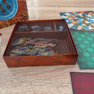 K&ouml;nnte beinhalten: Eine braune Brettspielbox mit einem Metallgitterdeckel, die Spielteile enth&auml;lt. Die Box steht auf einer Holzoberfl&auml;che, mit Spielbrettern und Anleitungen in der N&auml;he. Die Spielbretter weisen farbenfrohe Designs und Muster auf.
