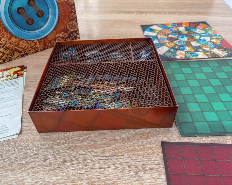 K&ouml;nnte beinhalten: Eine braune Brettspielbox mit einem Metallgitterdeckel, die Spielteile enth&auml;lt. Die Box steht auf einer Holzoberfl&auml;che, mit Spielbrettern und Anleitungen in der N&auml;he. Die Spielbretter weisen farbenfrohe Designs und Muster auf.