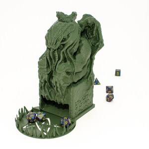 May include: Green 3D printed dice tower shaped like a Cthulhu monster with a dice tray at the bottom. The tower has a slot for dice to fall through and a small opening at the top for rolling dice. There are several blue and purple dice scattered around the tower.
