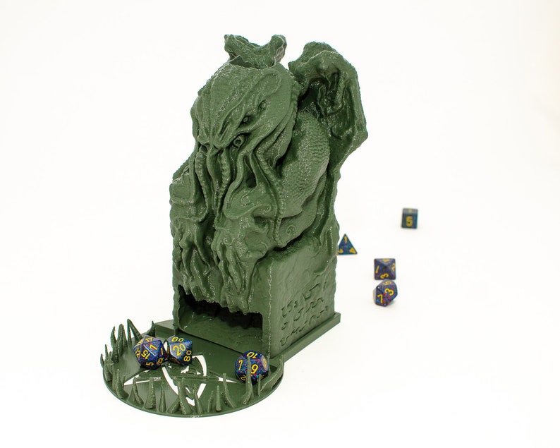May include: Green 3D printed dice tower shaped like a Cthulhu monster with a dice tray at the bottom. The tower has a slot for dice to fall through and a small opening at the top for rolling dice. There are several blue and purple dice scattered around the tower.