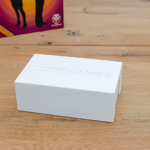 Op de afbeelding: Een witte doos met het woord "CODENAMES" erop gegraveerd. De doos staat op een houten oppervlak.