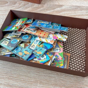 K&ouml;nnte beinhalten: Eine braune, rechteckige Schale gef&uuml;llt mit bunten Spielsteinen. Die Spielsteine weisen verschiedene Designs auf, darunter Kn&ouml;pfe, Fische und geometrische Muster. Die Schale steht auf einer hellen Holzoberfl&auml;che.