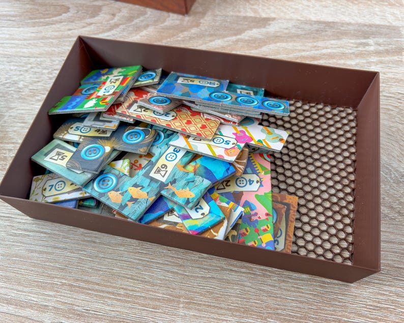 K&ouml;nnte beinhalten: Eine braune, rechteckige Schale gef&uuml;llt mit bunten Spielsteinen. Die Spielsteine weisen verschiedene Designs auf, darunter Kn&ouml;pfe, Fische und geometrische Muster. Die Schale steht auf einer hellen Holzoberfl&auml;che.