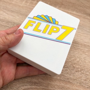 May include: A white rectangular game box with the text "FLIP 7" in yellow, blue, and purple. The box is held in a hand and rests on a wooden surface. The game box has a graphic design of a fan.
