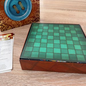 K&ouml;nnte beinhalten: Ein Brettspielkasten mit einem gr&uuml;n-schwarz karierten Spielbrett. Die Box ist braun mit einem dekorativen Design. Eine Spielanleitung ist links von der Box sichtbar. Die Oberseite der Box hat ein blaues Knopfdessign.