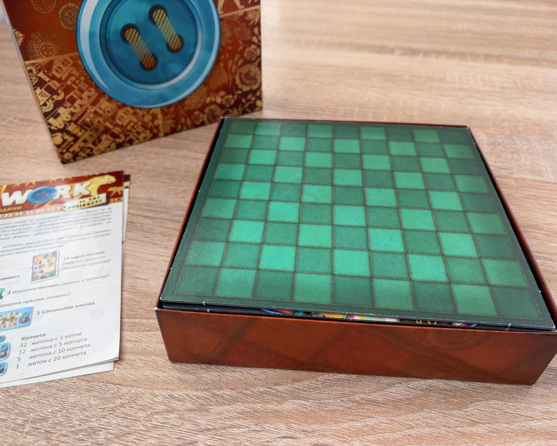 K&ouml;nnte beinhalten: Ein Brettspielkasten mit einem gr&uuml;n-schwarz karierten Spielbrett. Die Box ist braun mit einem dekorativen Design. Eine Spielanleitung ist links von der Box sichtbar. Die Oberseite der Box hat ein blaues Knopfdessign.