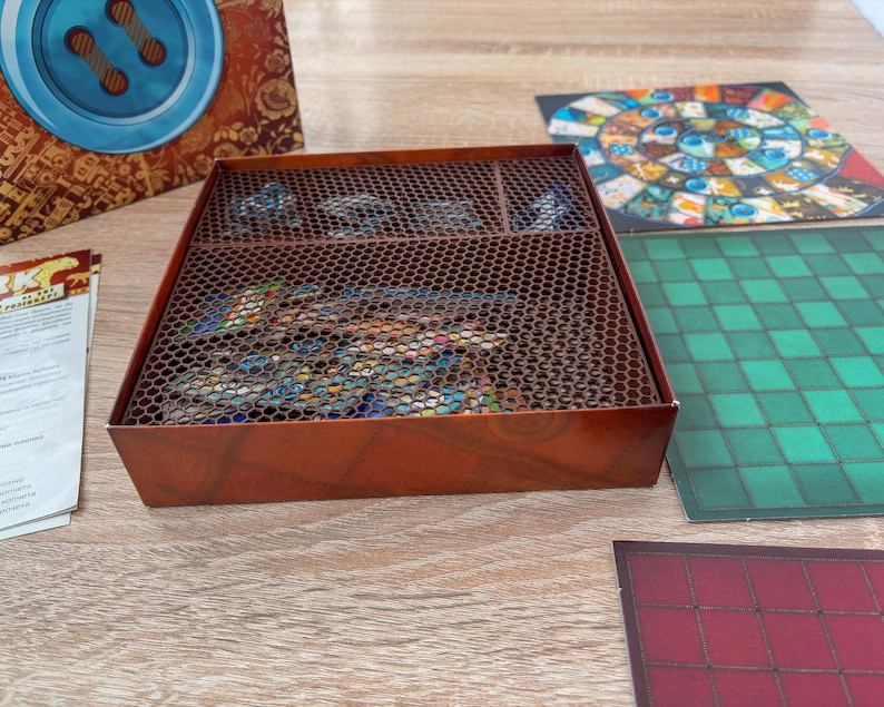 K&ouml;nnte beinhalten: Eine braune Brettspielbox mit einem Metallgitterdeckel, die Spielteile enth&auml;lt. Die Box steht auf einer Holzoberfl&auml;che, mit Spielbrettern und Anleitungen in der N&auml;he. Die Spielbretter weisen farbenfrohe Designs und Muster auf.