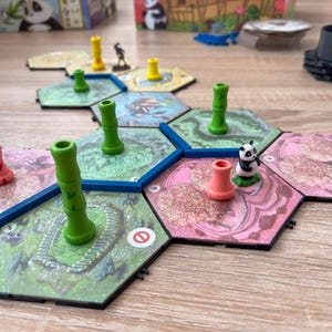 Peut inclure: Jeu de société avec des tuiles hexagonales représentant des tiges de bambou et une figurine de panda. Les tuiles sont vertes, roses et bleues, avec des pions jaunes et roses. Le jeu est posé sur une surface en bois brun clair.