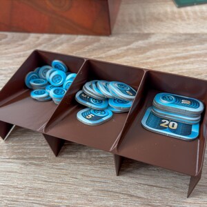 K&ouml;nnte beinhalten: Ein brauner Spiel-Organizer aus Kunststoff mit drei F&auml;chern, gef&uuml;llt mit blau-wei&szlig;en Spielmarken. Die Marken haben ein kreisf&ouml;rmiges Design und die Zahlen 5 und 20. Der Organizer befindet sich auf einer Holzoberfl&auml;che.