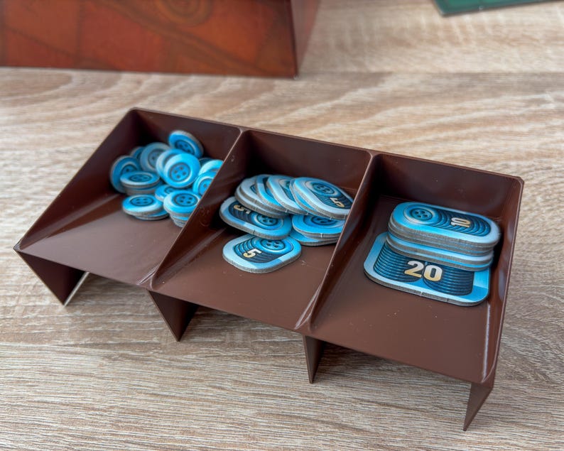 K&ouml;nnte beinhalten: Ein brauner Spiel-Organizer aus Kunststoff mit drei F&auml;chern, gef&uuml;llt mit blau-wei&szlig;en Spielmarken. Die Marken haben ein kreisf&ouml;rmiges Design und die Zahlen 5 und 20. Der Organizer befindet sich auf einer Holzoberfl&auml;che.