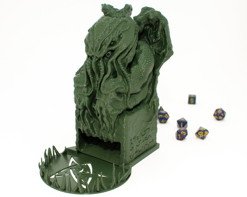 May include: Green 3D printed dice tower shaped like a Cthulhu monster with a circular base with spikes. The tower has a slot for dice to fall out of. There are several blue dice with yellow numbers on a white surface.
