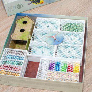May include: A white and blue game organizer with a birdhouse, a bird, and a variety of colorful compartments. The organizer is designed to hold game pieces and accessories.
