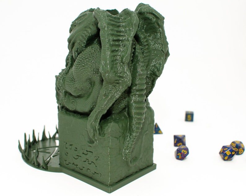 May include: Green 3D printed dragon dice tower with a circular base with spikes. The dragon is curled around a square base with runes carved into it. There are several blue and gold dice scattered around the tower.