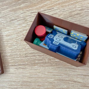 K&ouml;nnte beinhalten: Eine braune rechteckige Box enth&auml;lt verschiedene Holzspielsteine. Ein blaues, fahrzeugf&ouml;rmiges Holzteil ist prominent, zusammen mit roten und gr&uuml;nen kreisf&ouml;rmigen Teilen und kleinen rechteckigen Teilen mit gelben Punkten.