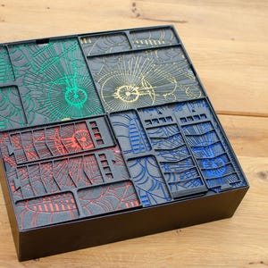 Puede incluir: Una caja negra con tapa abatible, que contiene cinco azulejos cuadrados con intrincados patrones en rojo, verde, azul y dorado. Los azulejos están dispuestos en forma de cuadrícula.