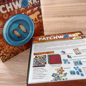 K&ouml;nnte beinhalten: Das Brettspiel "Patchwork" wird gezeigt, mit seiner Schachtel, die eine gro&szlig;e Knopfgrafik und den Titel des Spiels zeigt. Die Schachtel ist braun mit goldenen Akzenten. Spielkomponenten sind sichtbar, darunter Spielbretter, Spielsteine und Karten.
