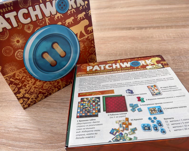 K&ouml;nnte beinhalten: Das Brettspiel "Patchwork" wird gezeigt, mit seiner Schachtel, die eine gro&szlig;e Knopfgrafik und den Titel des Spiels zeigt. Die Schachtel ist braun mit goldenen Akzenten. Spielkomponenten sind sichtbar, darunter Spielbretter, Spielsteine und Karten.