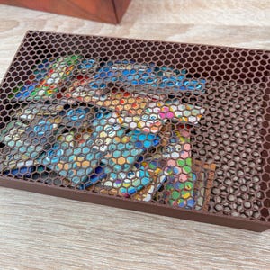 K&ouml;nnte beinhalten: Eine braune, rechteckige Box mit einem Deckel im Wabenmuster. Sie enth&auml;lt bunte, gemusterte Gegenst&auml;nde, die teilweise durch den Deckel sichtbar sind. Die Box besteht aus einem dunkelbraunen Material und steht auf einer hellen Holzoberfl&auml;che.
