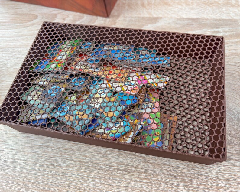 K&ouml;nnte beinhalten: Eine braune, rechteckige Box mit einem Deckel im Wabenmuster. Sie enth&auml;lt bunte, gemusterte Gegenst&auml;nde, die teilweise durch den Deckel sichtbar sind. Die Box besteht aus einem dunkelbraunen Material und steht auf einer hellen Holzoberfl&auml;che.