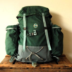 Könnte beinhalten: Ein dunkelgrüner Rucksack mit zwei großen Seitentaschen, hellgrauen verstellbaren Riemen und einem grauen Bodenbereich mit Kordelzug und geknoteter Schnur. Ein kleines Logo-Patch mit dem Schriftzug "walker" befindet sich mittig auf der Vorderseite.