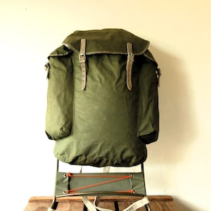 May include: An olive green canvas external frame backpack featuring a large main compartment with a top flap secured by two light-coloured leather straps with metal buckles, two side pockets, and a metal frame base with orange cord lacing.
