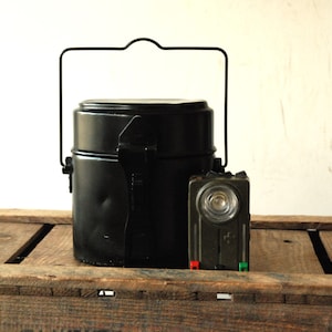 Op de afbeelding: Een zwarte metalen campingkookset met een draaggreep en een zwarte zaklamp met een witte lens.