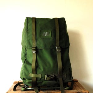 Può includere: Uno zaino verde bosco dotato di una patta superiore con due cinghie verticali in nylon verde oliva e fibbie in metallo. La parte anteriore della patta include una piccola finestra rettangolare in plastica trasparente per l'identificazione.