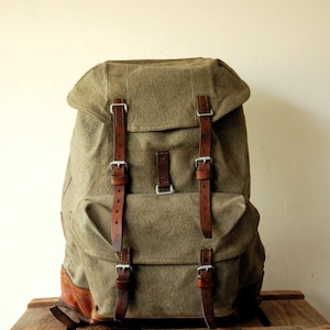 Puede incluir: Una mochila de lona color verde oliva con adornos y correas de cuero marrón. La bolsa incluye una solapa superior con dos correas verticales de cuero, un bolsillo frontal con dos correas adicionales y hebillas de metal plateado para el cierre.