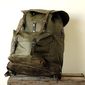 Può includere: Uno zaino in tela verde oliva con un ampio scomparto principale con patta, una tasca frontale e cinghie in pelle marrone scuro con fibbie in metallo. La base dello zaino è rinforzata con un pannello in pelle marrone scuro.
