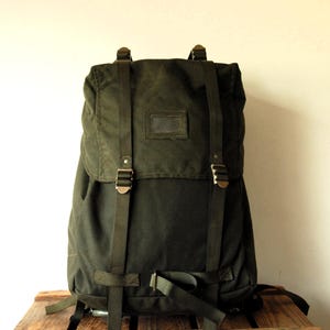 Op de afbeelding: Een donker olijfgroene canvas rugzak met een bovenflap voorzien van twee verticale verstelbare nylon banden en metalen gespen. De voorkant van de flap bevat een rechthoekig venster voor identificatie. De tas heeft een gestructureerd ontwerp.