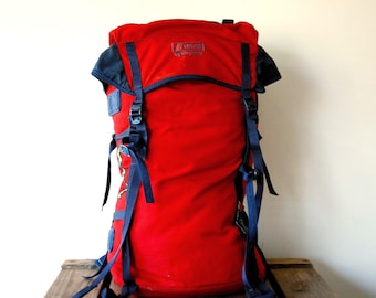 May include: A red backpack with dark blue straps and buckles. The front features the text "VROTO by berghaus" in white lettering. The bag has a top flap closure, side compression straps, and additional attachment points on the sides.