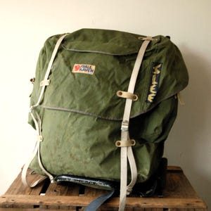 Op de afbeelding: Een olijfgroene Fjällräven-rugzak met een grote klep aan de bovenkant, twee verticale beige banden met metalen gespen en een zwarte zijpatch met de gele tekst "MILE". De tas heeft een stevige framebasis en een rechthoekig logoplaatje.
