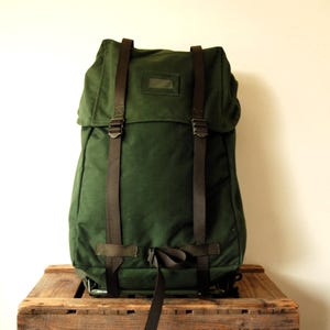 Può includere: Uno zaino in tela verde scuro con un'ampia patta superiore, due cinghie verticali in tessuto marrone con fibbie regolabili e una piccola finestra rettangolare per l'identificazione sulla patta frontale. Design strutturato con finiture marroni.