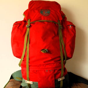Könnte beinhalten: Ein leuchtend roter Rucksack mit olivgrünen Riemen, seitlichen Reißverschlusstaschen und einem runden Aufnäher auf der oberen Klappe mit dem Text Outdoor Leisure. Der Boden des Rucksacks ist olivgrün und verfügt über Metallbeschläge und verstellbare Riemen.
