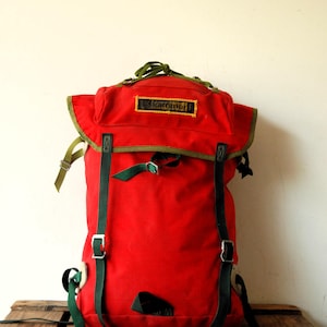 Peut inclure: Un sac à dos rouge vif Karrimor doté de sangles en nylon vert foncé, de boucles métalliques et d'un rabat supérieur. Le sac présente un écusson rectangulaire avec le texte Karrimor cousu sur le rabat avant, rehaussé d'une bordure vert olive.