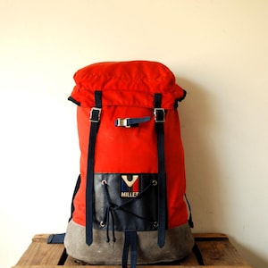 Puede incluir: Una mochila de color rojo brillante con un bolsillo frontal de aspecto cuero azul oscuro con detalle de cordones y el logotipo de Millet. Incluye correas de nailon azul oscuro con hebillas metálicas, cierre de solapa y base de ante gris.