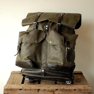 May include: Olive green backpack with black leather accents. Features multiple pockets, adjustable straps, and metal buckles. The bag is made of a durable, water-resistant material, ideal for outdoor adventures.