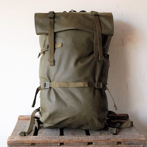Könnte beinhalten: Ein olivgrüner Rolltop-Rucksack mit verstellbaren Nylongurten und Kunststoffschnallen. Die Tasche hat ein vertikales Design mit einem Rollverschluss, der durch zwei vordere Riemen und einen horizontalen Kompressionsriemen in der Mitte gesichert ist.