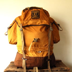 Può includere: Uno zaino Karrimor color giallo senape e marrone con una patta superiore dotata di una toppa nera con la scritta Karrimor e Jaguar IV. Lo zaino include cinghie beige regolabili, fibbie in metallo, tasche laterali e uno scomparto con cerniera alla base.