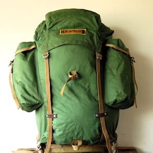 Könnte beinhalten: Ein grüner Karrimor-Rucksack mit zwei großen Seitentaschen, einem Deckelfach und beigen Gurtbändern mit Metallschnallen. Auf der Vorderseite befindet sich ein gelb-schwarzes Karrimor-Markenetikett. Der Rucksack ist auf einem silbernen externen Metallrahmen montiert.