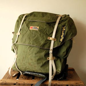 Könnte beinhalten: Ein olivgrüner Fjällräven-Rucksack mit großer Deckelklappe, zwei vertikalen beigen Riemen mit Metallschnallen und einem schwarzen Aufnäher an der Seite mit dem gelben Schriftzug "MILE". Der Rucksack hat einen Rahmenboden und ein Logo-Patch.
