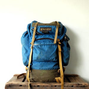 Könnte beinhalten: Ein blauer und olivgrüner Karrimor-Rucksack mit oberer Klappe, einer zentralen Vordertasche und zwei Seitenfächern. Der Rucksack verfügt über lange beigefarbene Riemen mit Metallschnallen und ein Karrimor-Markenetikett im oberen Bereich.
