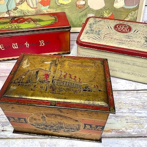 May include: Three vintage tin boxes with colourful designs. The top box is red with gold trim and the text "East West Homes Best". The middle box is gold with red trim and a scene of a building with flags. The bottom box is white with red trim and the text "1953 Coronation" and "By the Grace of God the Faith Elizabeth".
