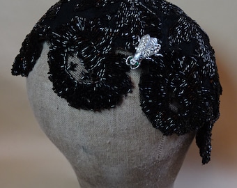 Puede incluir: Un sombrero negro de cuentas con un broche de abeja de plata decorativo. La abeja tiene alas verdes y diamantes de imitación transparentes.